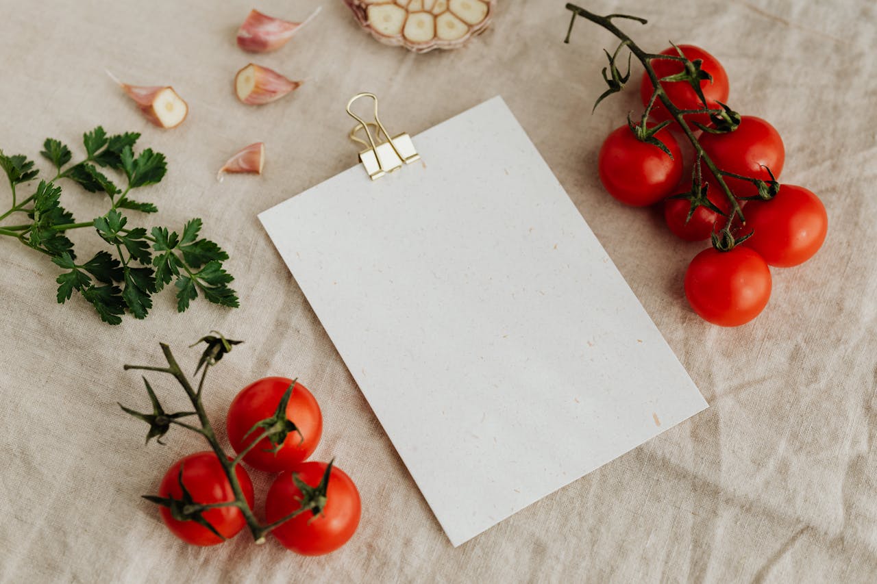 services-02 Top view of blank clipboard with golden paper binder placed on linen tablecloth among tasty red tomatoes on branches together with chopped garlic head and green parsley ideal for recipe or menu